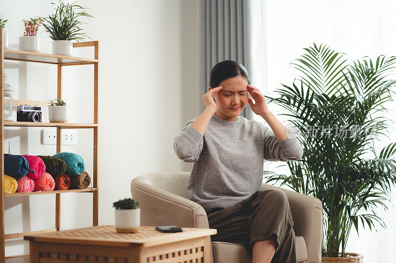 一名头痛的亚洲妇女坐在家里客厅的扶手椅上。