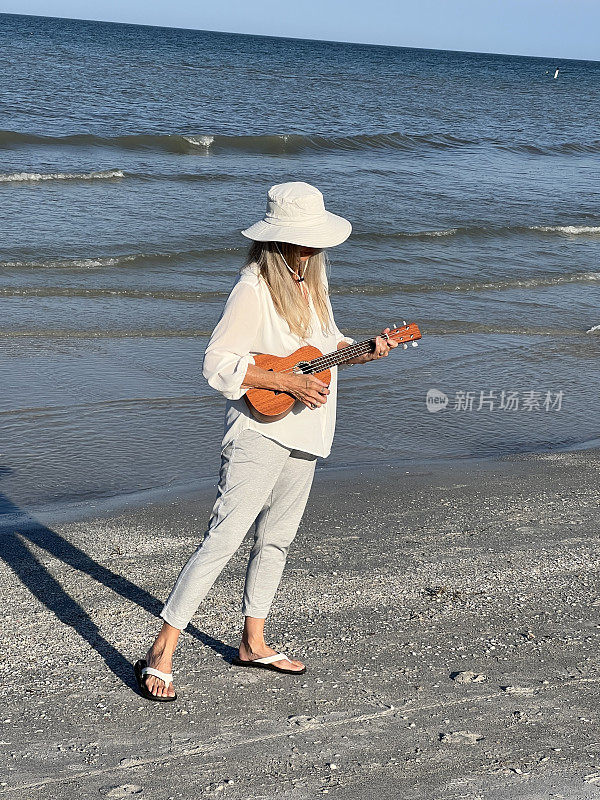 一位老年妇女走在佛罗里达海滩上，对着水和天空弹奏着尤克里里