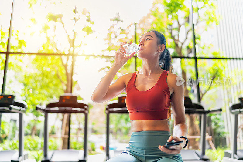 亚洲女性在阳光灿烂的室内健身俱乐部喝着水，一边使用智能手机，一边休息