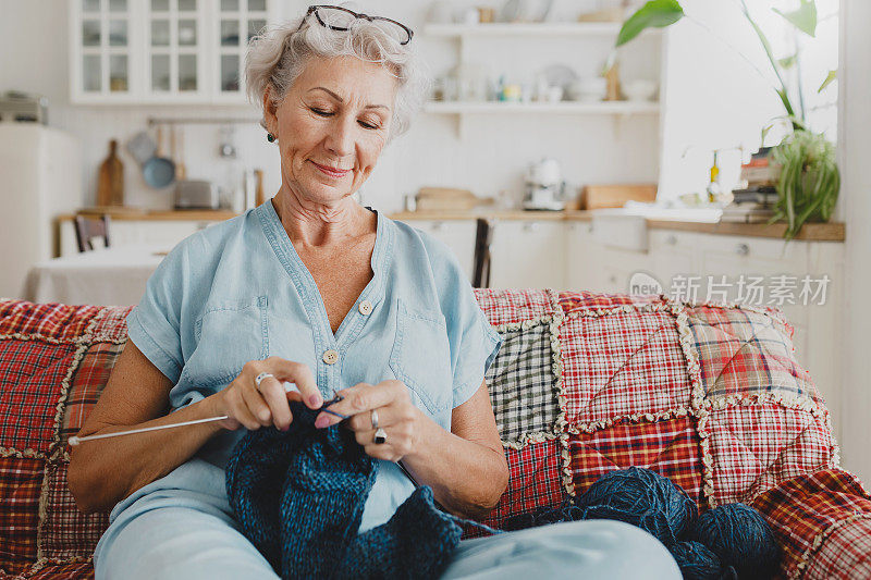 快乐的奶奶，花白的头发，时尚的衣服，享受着为她的丈夫织温暖的毛衣，或为她的孙子织时尚的围巾，坐在沙发上，用蓝色的纱线球，熟练地拿着针