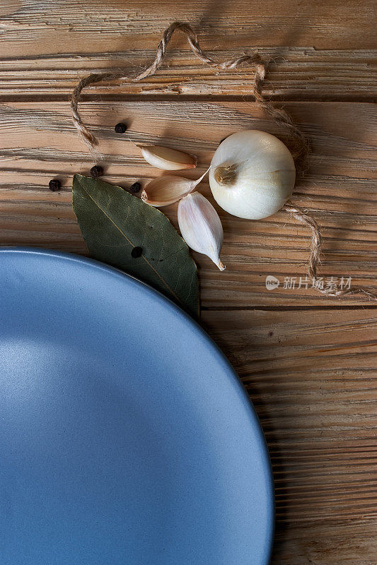 洋葱，大蒜，黑胡椒和月桂叶与空的蓝色盘子在木背景