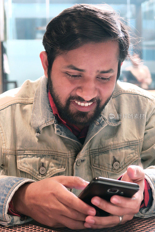 这是一个年轻的印度人，印地语血统，微笑着慢慢开始新的一天，阅读手机短信，浏览早间互联网报纸，在智能手机上嘲笑社交媒体，留着黑胡子，穿着磨砂蓝色牛仔夹克