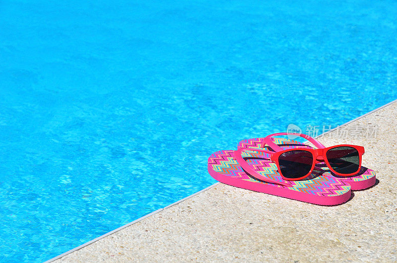 蓝色的游泳池旁边是红色的人字拖和太阳镜