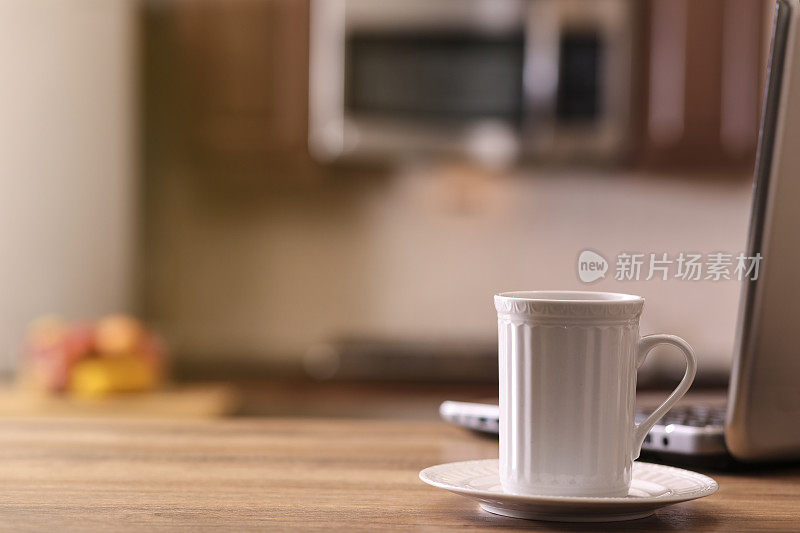 离焦厨房，咖啡杯，笔记本电脑在木头桌子的前景。