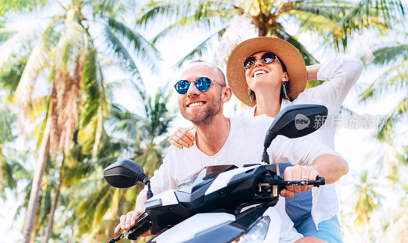 快乐的微笑旅行者夫妇骑摩托车在他们的热带度假棕榈树下