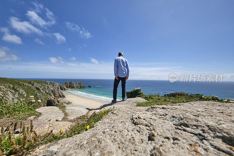 一个人站在一块岩石上，看着佩顿·范德海滩到洛根的岩石，在一个完美的六月的下午，康沃尔。