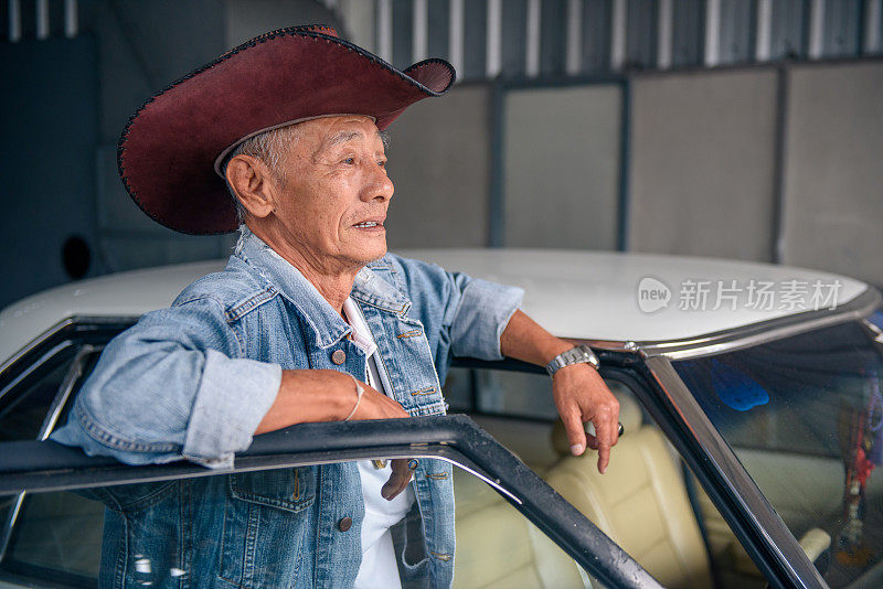 年长的亚洲男人在复古牛仔夹克摆姿势与古董车