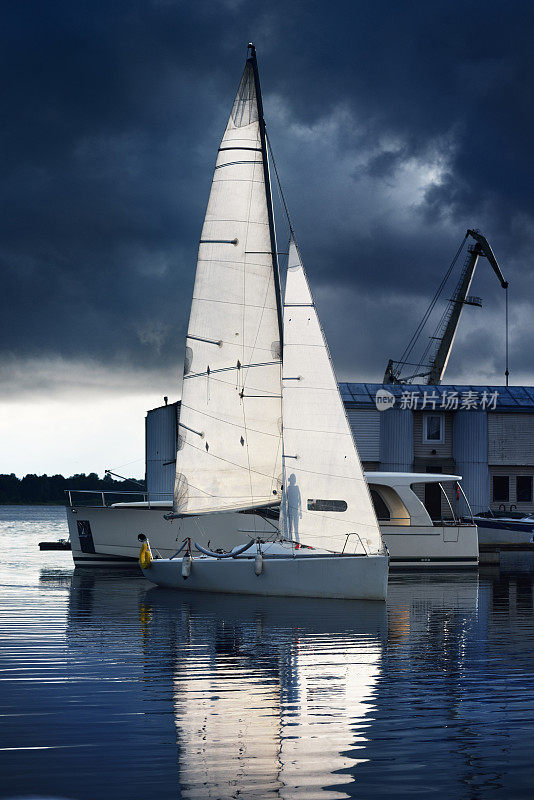 现代小型24英尺帆船赛帆船在日落时停泊在码头上。女孩在船帆后面剪影。黑暗风暴的天空。拉脱维亚的里加港