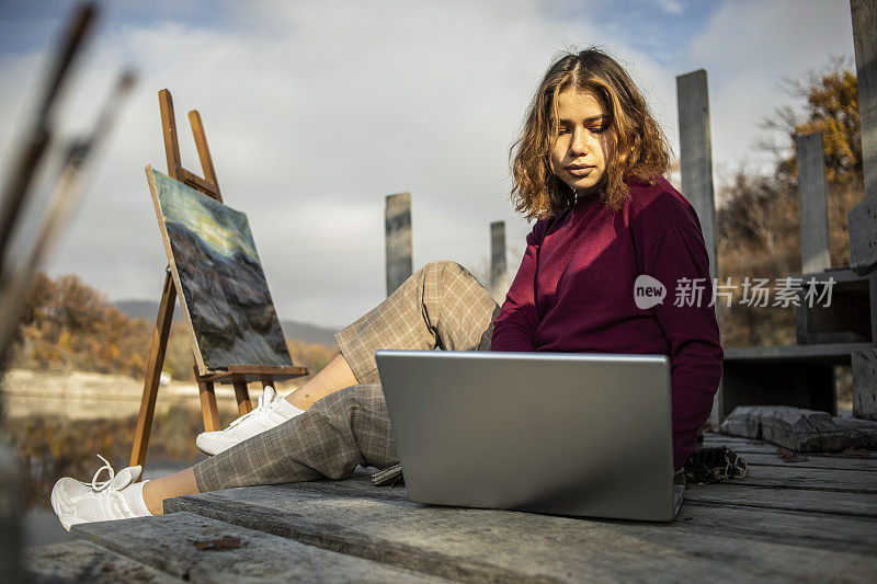 这位年轻的艺术家正在湖边休息，并在她的笔记本电脑上寻找新的绘画技法——女艺术家户外绘画