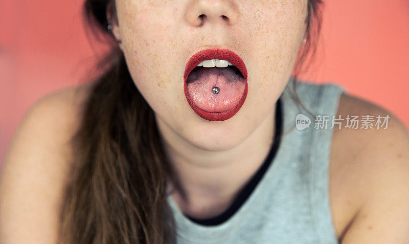 近距离肖像的年轻女子与雀斑突出穿舌，显示她的舌穿孔与粉红色的背景
