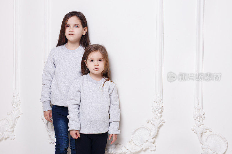 两个可爱的小女孩，穿着毛衣和牛仔裤，在白色背景下一起拍照。空间复制。