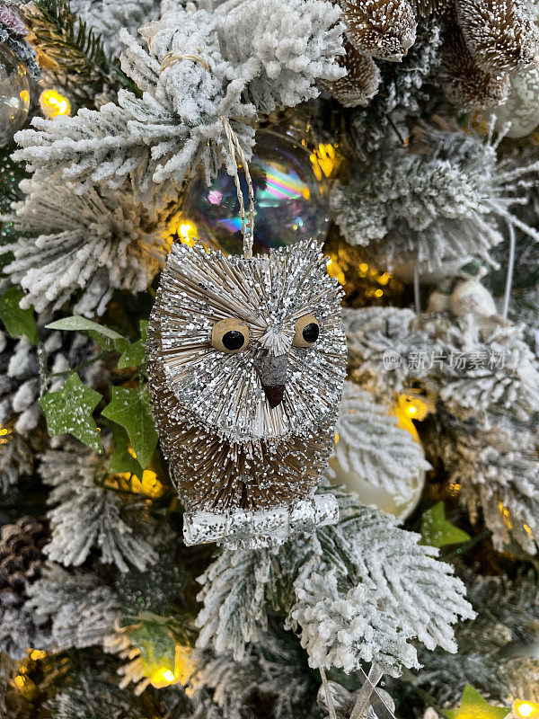 特写图像猫头鹰装饰松果在人造圣诞树的树枝上，云杉针覆盖着人工喷雾雪，模糊的白色精灵灯的背景