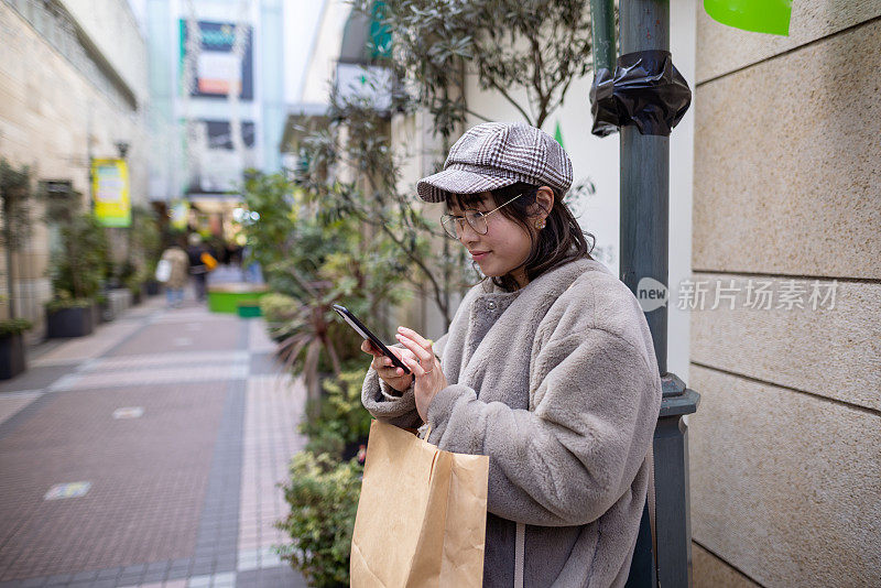 快乐的女人靠在柱子上，在购物街上使用智能手机