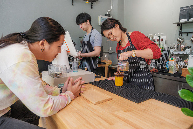 年轻的亚洲咖啡师或咖啡店老板在咖啡馆的吧台为顾客制作一杯冰橙咖啡