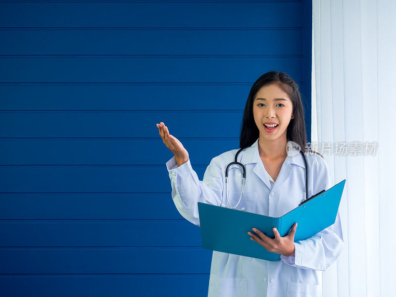 微笑美丽的亚洲女医生画像站在蓝色背景，拿着文件文件夹，并在空白上显示手势。自信的年轻女性从业人员拿着文件文件夹。