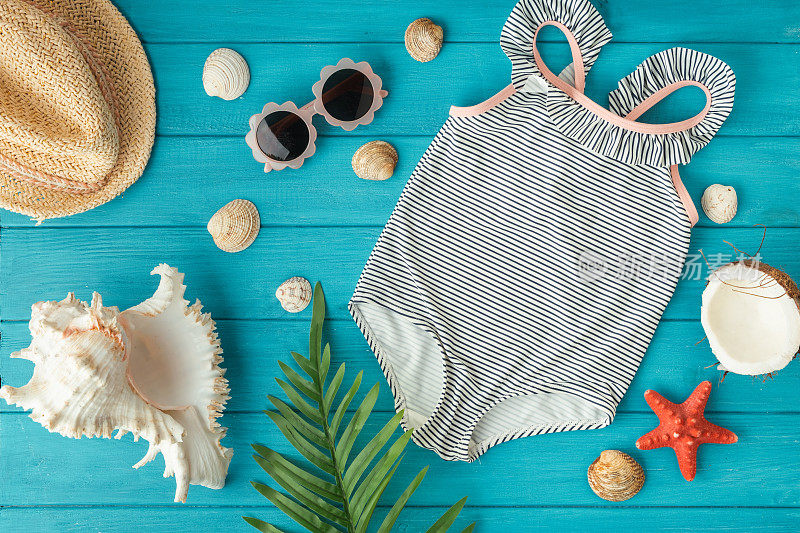 孩子们连体泳衣和草帽旁边的海滩配件，贝壳，海星和椰子在木蓝绿色的背景。夏日氛围和海边度假的概念。平躺