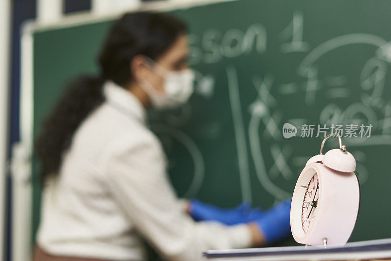 桌子上的闹钟，背景是一位戴着口罩正在讲课的女老师