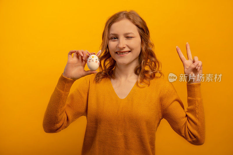 快乐的微笑眨眼的女孩拿着白色的鸡蛋装饰粉红色的兔子耳朵，花和兔子的口吻和显示和平手势v标志手孤立在黄色背景。复活节的概念。