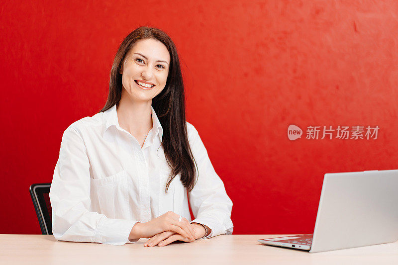 在红墙的办公室里，一位穿白衬衫的褐发女子在电脑前工作。