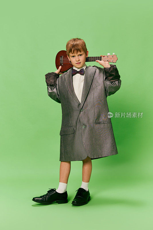 时尚的小学龄男孩在巨大的超大复古风格的衣服和大的男性鞋摆姿势与尤克里里吉他孤立在绿色背景。