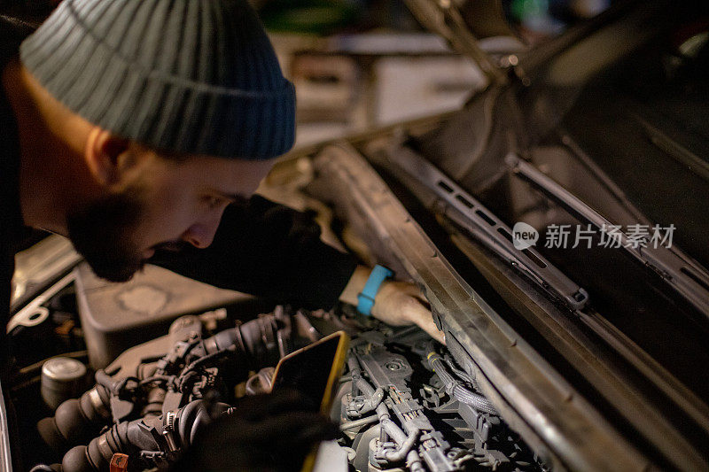 年轻男子在他的车库里自己维修汽车。他正在用手机给车钩点火。侧视图。