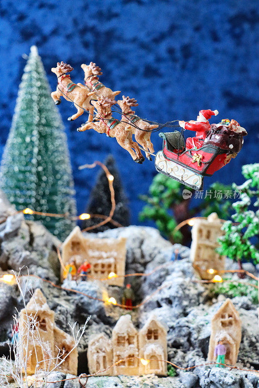 特写图像的圣诞老人雪橇和驯鹿飞过风景优美的圣诞村显示，人工塑料和木制圣诞树在山上设置，照明自制木屋，重点在前景