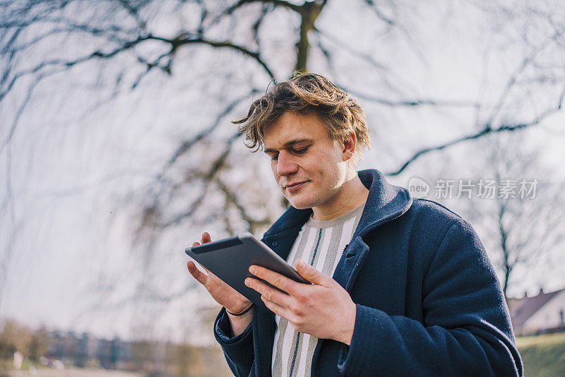 一个年轻人在河边的公园外使用数码平板电脑的正面照片。副本的空间。库存图片