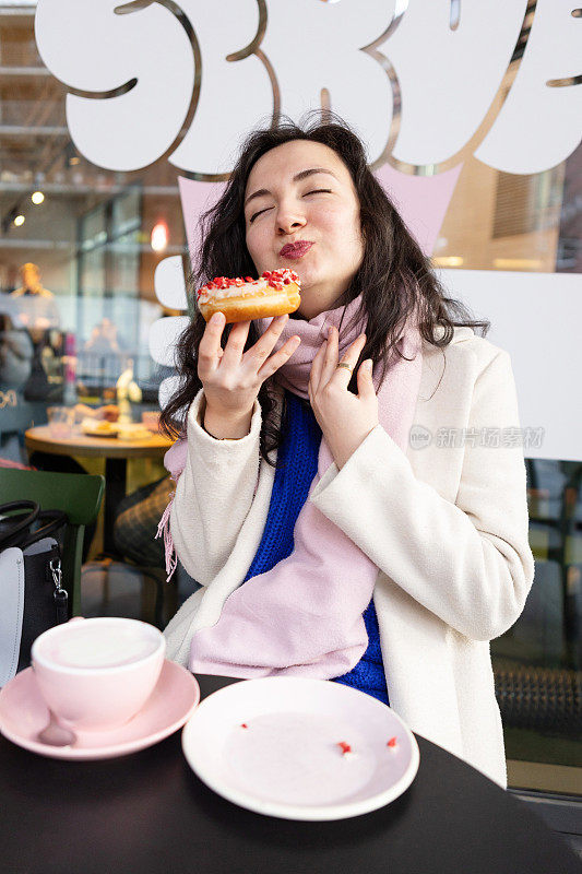 年轻的黑发女子穿着五颜六色的衣服，在咖啡馆里吃着甜甜圈，喝着粉红色的咖啡
