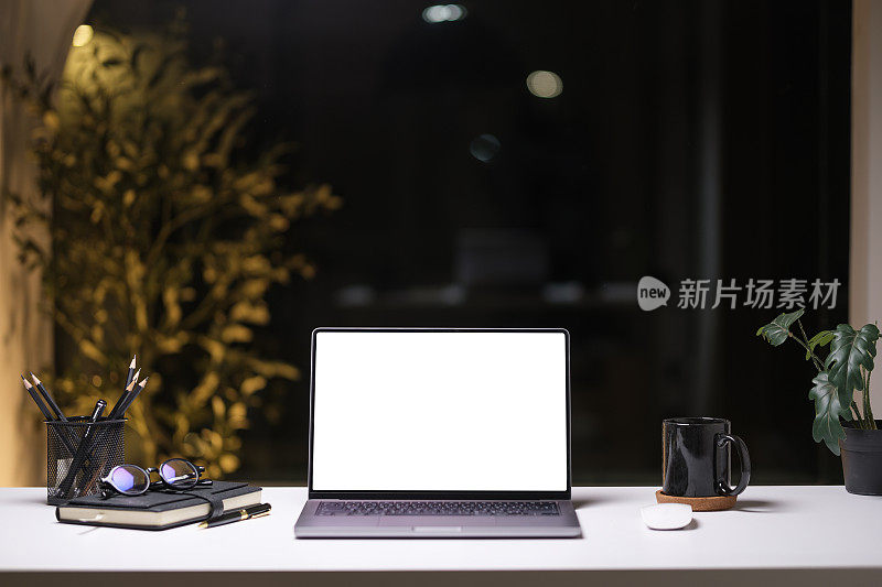 笔记本电脑与空白屏幕，咖啡杯和盆栽在白色的桌子上在现代黑暗的办公室晚上。