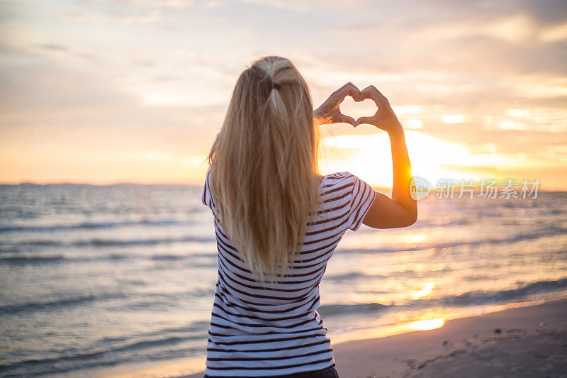 日落时分，沙滩上的女孩用手做出心形。