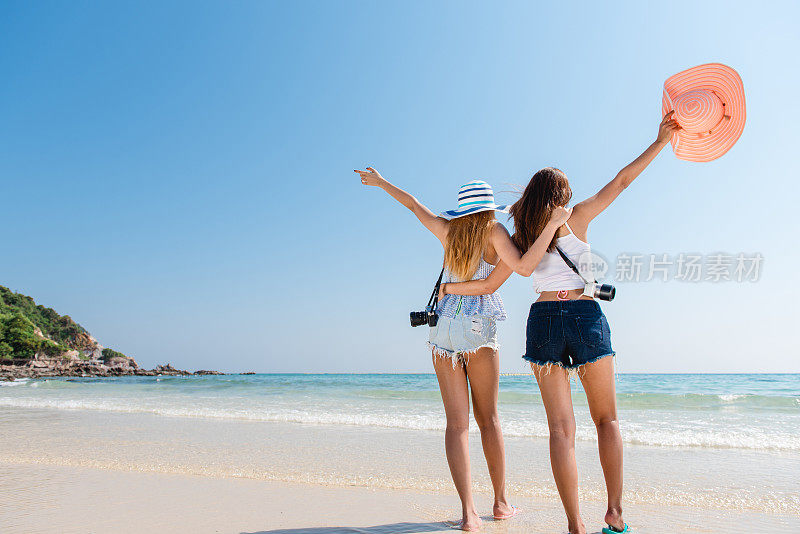 肖像两个年轻的亚洲女性朋友走在海边转身对着镜头笑。多种族的年轻女子在海滩上散步。