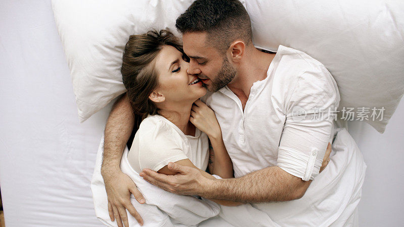 年轻美丽和相爱的夫妇亲吻和拥抱在床上，而早上醒来。俯视图有吸引力的男人亲吻和交谈他微笑的妻子