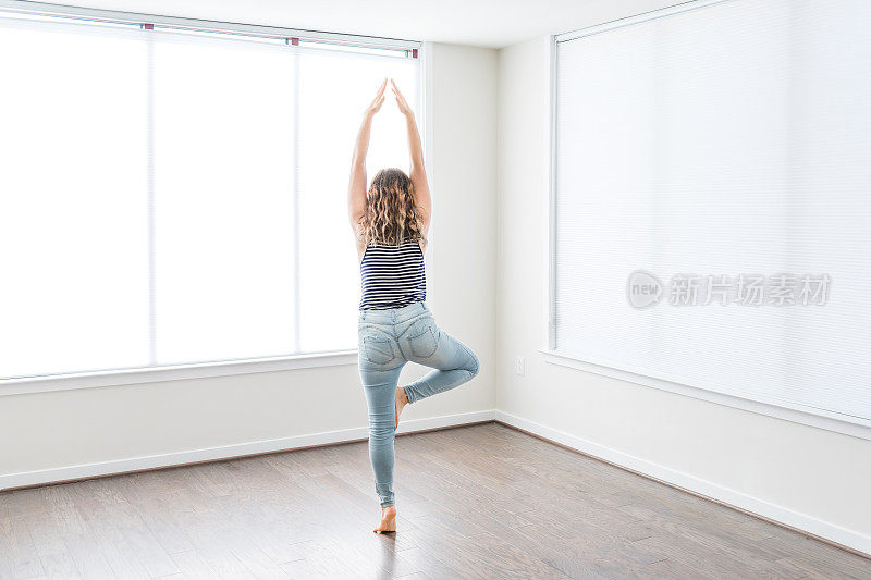年轻女子在树瑜伽姿势单腿站在空现代的新房间，硬木地板，大阳光窗户公寓，房子