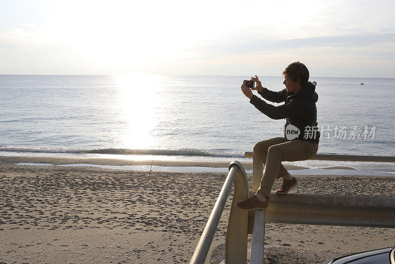 日出时分，一名年轻人在海边用智能手机拍照