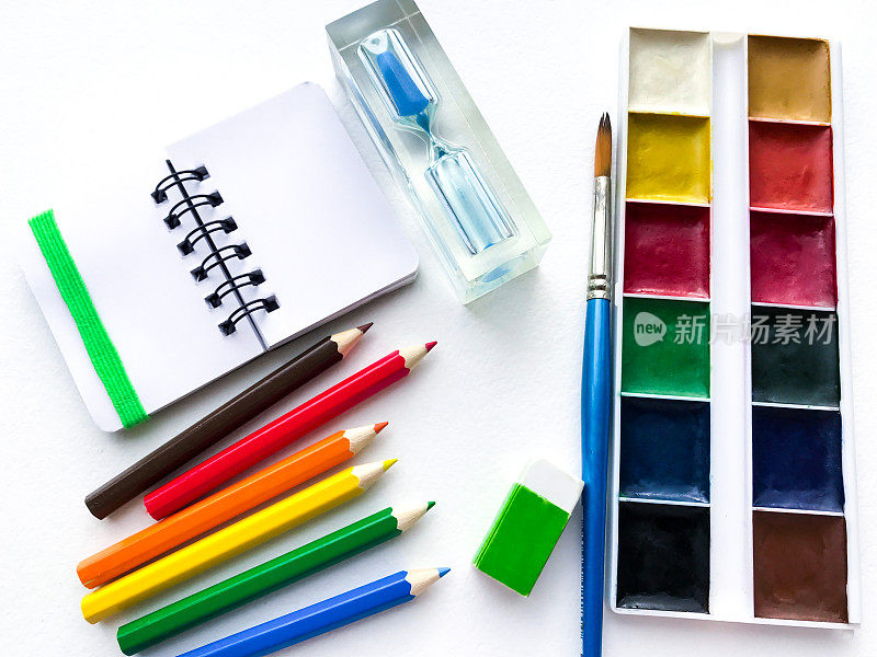 平铺文具学校和创意，绘画和工艺品(水彩画颜料，彩色铅笔，笔记本，沙漏)是在右边的白色水彩纸