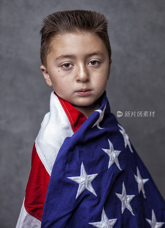 亚裔白人混血小男孩，浅黑色平顶发型，裹着美国国旗，在灰色的背景下摆姿势