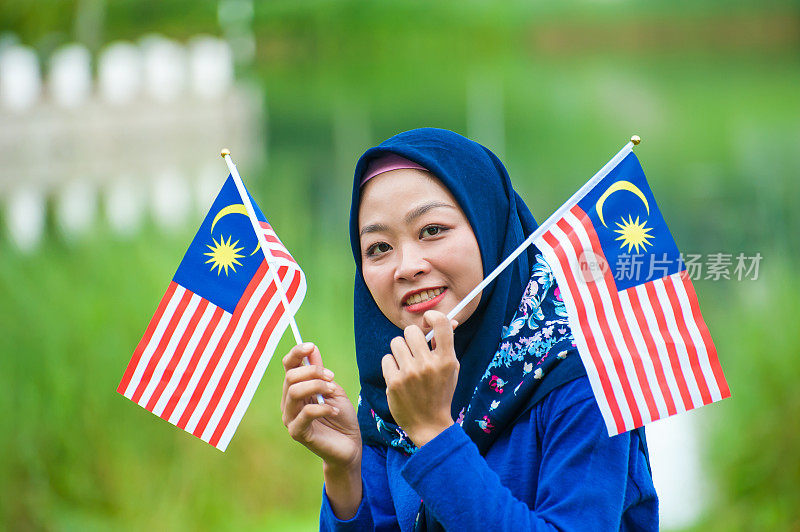 年轻的穆斯林妇女戴着头巾，挥舞着马来西亚国旗，具有高度的爱国主义精神，在社区花园庆祝独立日