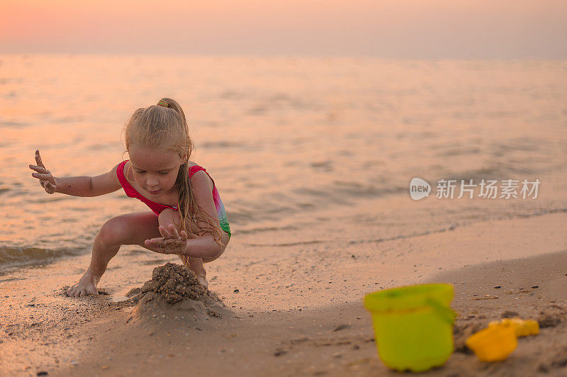 在夏天的黄金时间，傍晚日落时，金发小女孩在海边建了一座沙堡