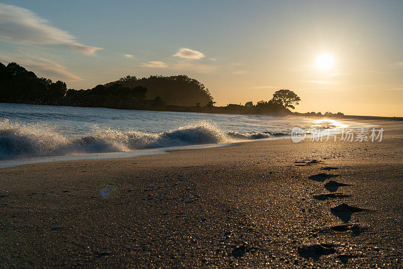 金色的日出在海岸特征脚印在沙子领先和汇聚到水的边缘在芒加努伊山新西兰。