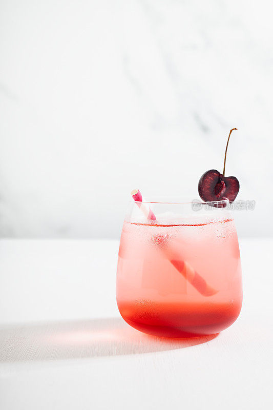 清凉的夏季饮料与樱桃利口酒在玻璃杯，切片水果，吸管，冰块在优雅阳光白色室内木桌子，大理石墙壁，垂直。