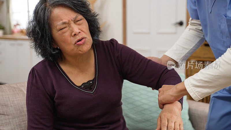 退休的亚洲女性病人呻吟痛苦的面部表情的特写，而护理助理是举起她的手臂做康复训练在家里