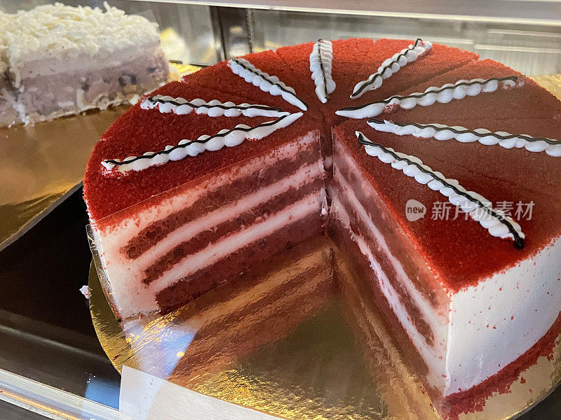 面包店展示甜点的特写图像，圆形的红色丝绒蛋糕切片，银色的蛋糕板，奶油糖霜分层海绵蛋糕片，管道糖霜装饰，重点放在前景