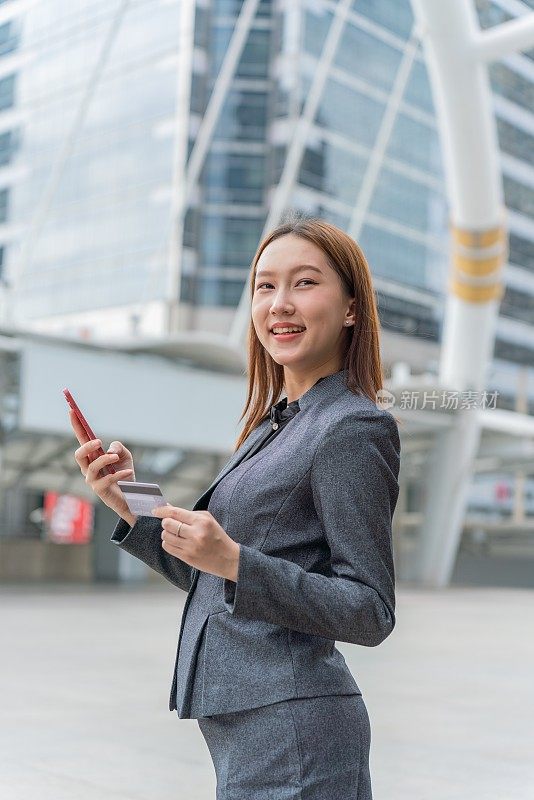 一名年轻的亚洲女商人穿着灰色的办公套装，在商业区用她的智能手机使用信用卡进行在线交易，她微笑着自信