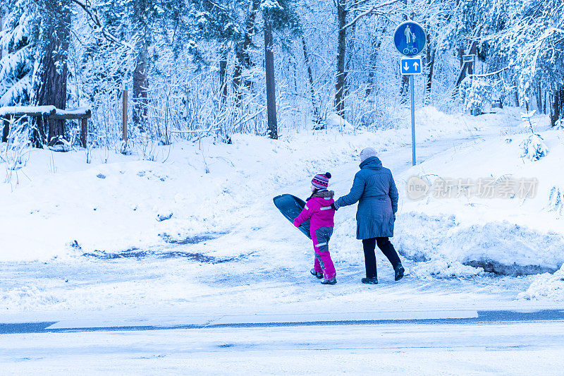 一位老妇人带着一个孩子在人行横道上穿过一条被雪覆盖的道路。