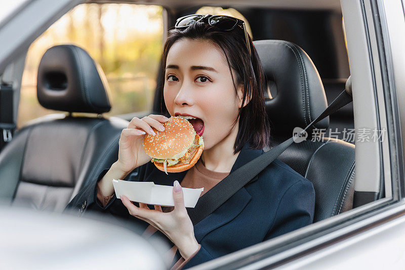 亚洲女人在车里休息，吃汉堡。一个年轻成功快乐的女人正在开车。