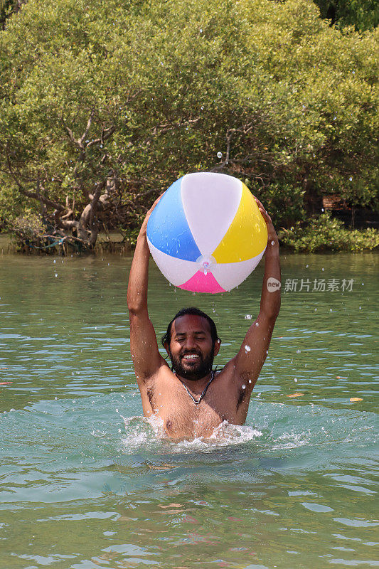 印度男子站在齐胸深的泻湖平静的水中，头顶上拿着彩色的塑料沙滩球，看着镜头，水溅起，看到有棕榈树的林地，前景聚焦
