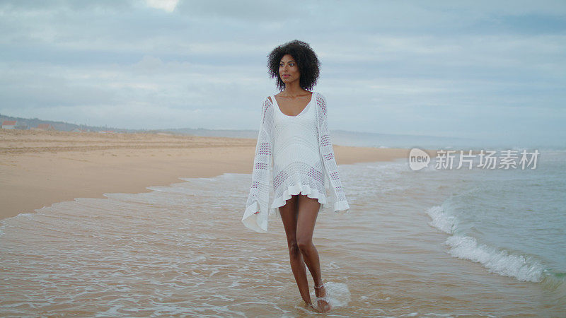 穿着白色衣服走在海岸线上的梦幻女人。体贴的卷发女孩去沙滩