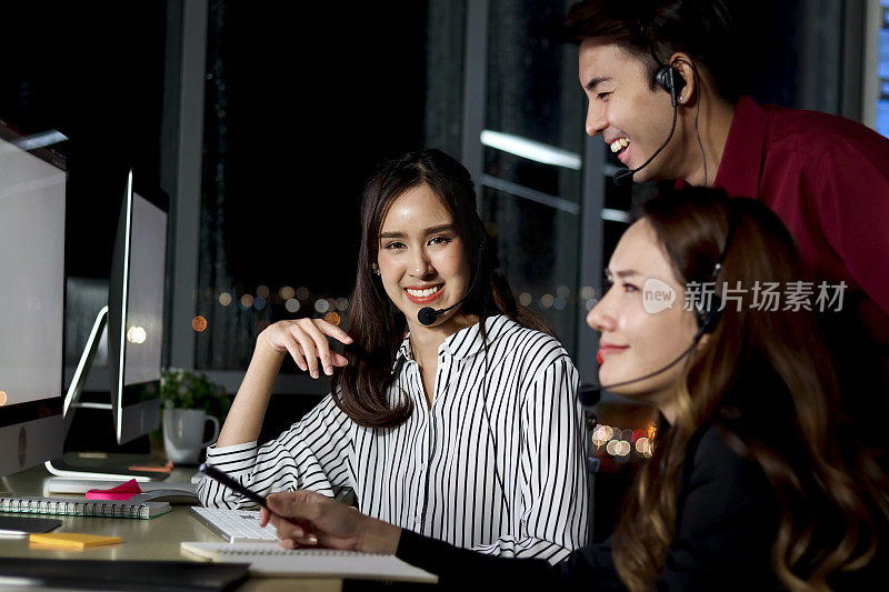 一群带耳机的亚洲夜班员工一起看着台式电脑讨论，一群带免提电话的夜班员工在呼叫中心客服关怀加班加点
