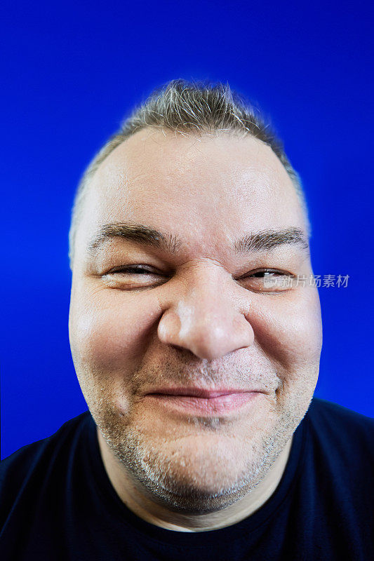 47岁白人男子的恶意微笑，肥胖问题，他的脸被蓝色背景的广角镜头扭曲，特写。