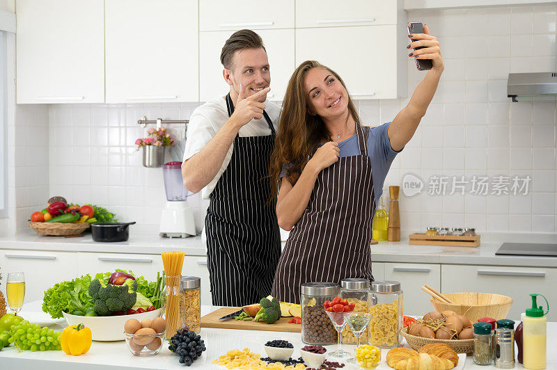 年轻情侣在家里的厨房里一起做饭。幸福的情侣用智能手机自拍或打视频电话。